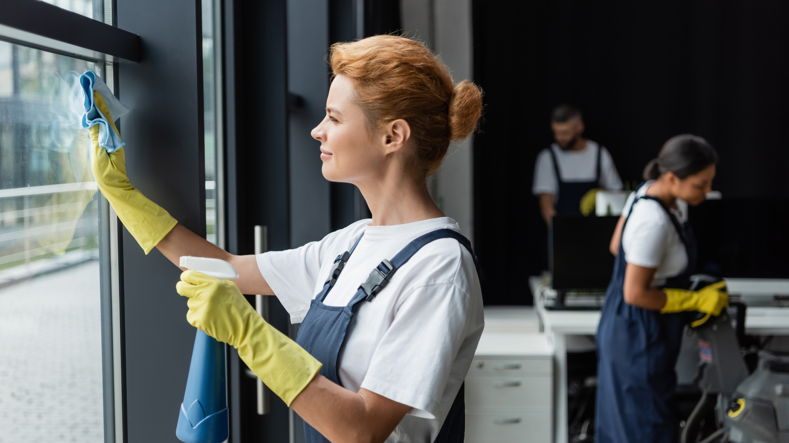 A person wearing a white shirt and blue overalls, with yellow rubber gloves, is cleaning a glass window with a cloth. In the background, two other individuals are engaged in cleaning tasks in an office environment.
